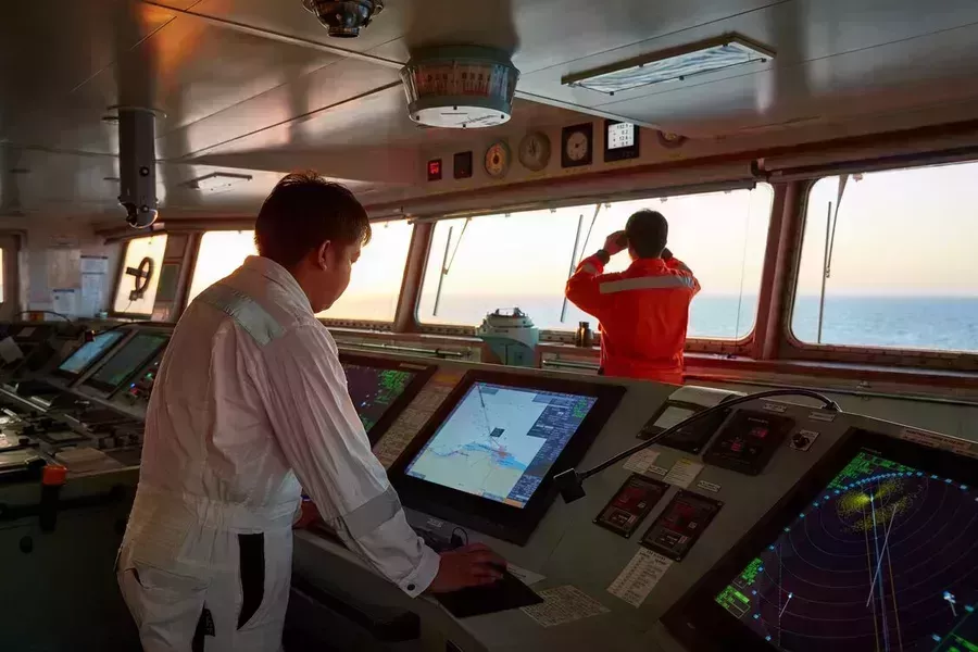 Two mariners on vessel, one looking at an ECDIS screen, one looking out to sea
