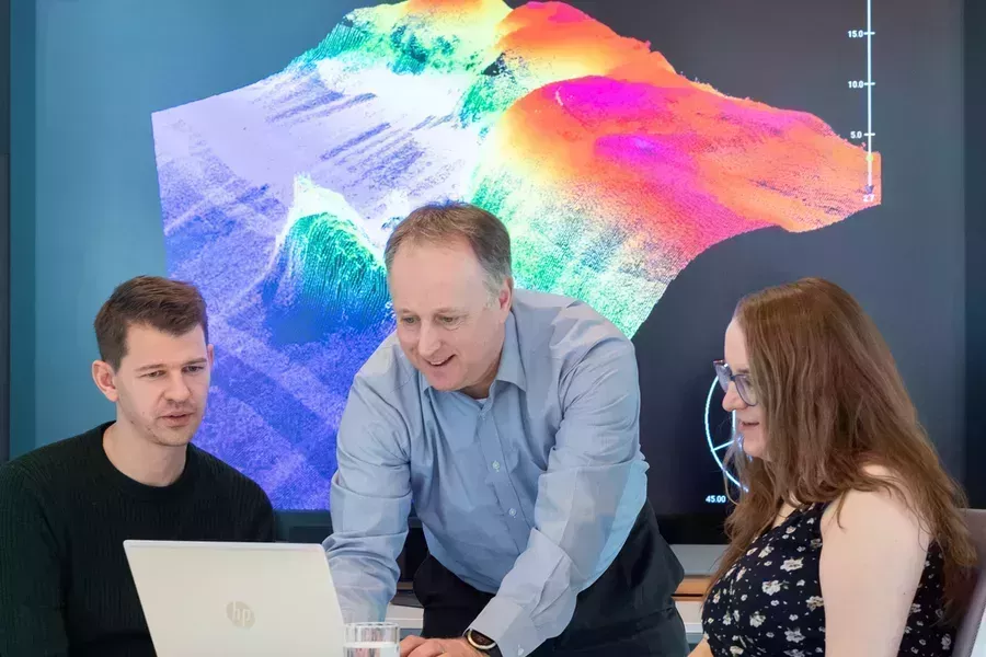 Colleagues gathered around a laptop in front of a screen displaying bathymetric data