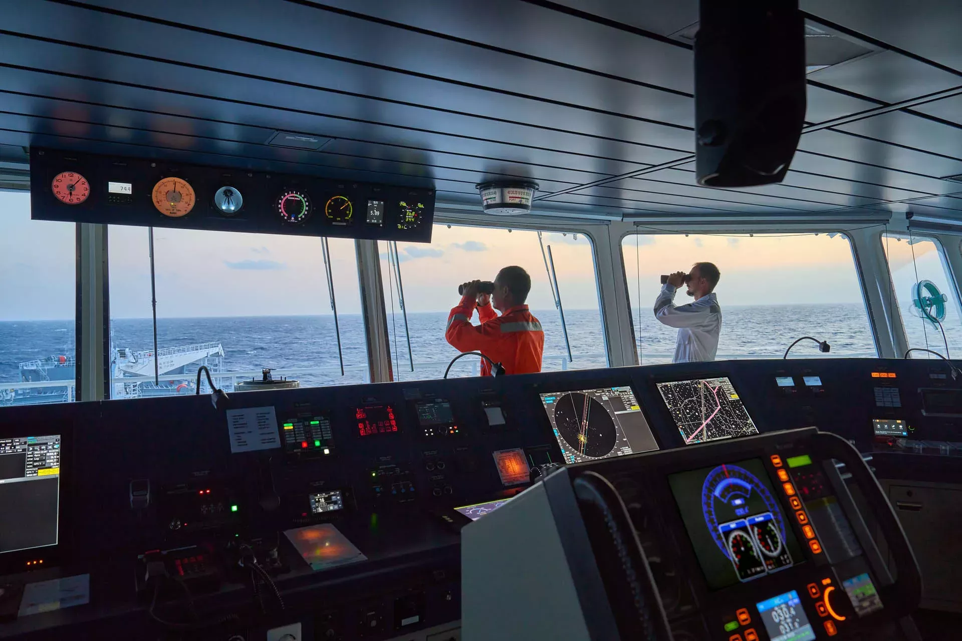 Two mariners on a ship's bridge looking out at sea with binoculars 